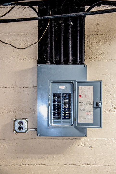 Stainless steel electrical circuit box open with circuits named by hand in basement on rough concrete block wall with conduit and ceiling and other pipes overhead painted black with dust on them Electrical Panel in a home