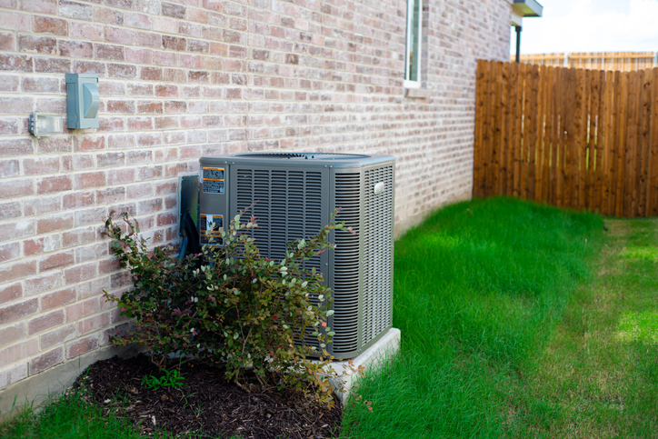 Outdoor HVAC unit and wall mounted electrical boxes installed alongside modern brick home, clean siding and windows, green grass landscaping shrub, privacy fence, disconnect switch, utility meter Condenser unit outside of house.